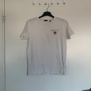 Vit t-shirt från GANT - Säljer en klassisk vit t-shirt från GANT med korta ärmar och rund hals. T-shirten har en liten logotyp på bröstet i form av en sköld med texten 'GANT'. Perfekt för en avslappnad stil.
