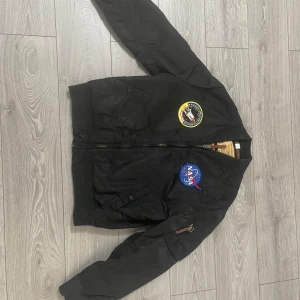 Svart bomberjacka med NASA-märken - Cool svart bomberjacka från Alpha Industries med NASA-märken. Jackan har dragkedja och ribbade muddar vid ärmslut och nederkant. Perfekt för dig som gillar rymdtema och stilren design. 🛰️