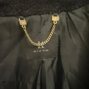 Svart kappa från Michaa - Elegant svart kappa från Michaa med klassisk design. Den har en stilren krage och långa ärmar, perfekt för kyligare dagar. Kappan är tillverkad i ett mjukt och hållbart material så den håller i flera år.