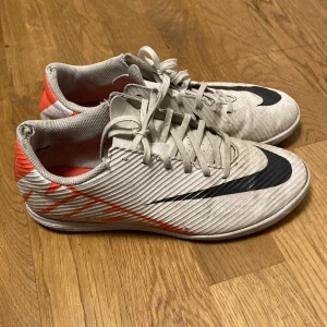 Vita och röda fotbollsskor från Nike - Säljer ett par vita fotbollsskor från Nike med svarta detaljer och röda accenter. Skorna har snörning och en strömlinjeformad design som passar perfekt för inomhusspel. De har en slitstark sula och är designade för att ge bra grepp på planen. Väldigt slitna och pris kan diskuteras. 