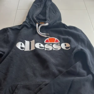 Svart hoodie från Ellesse - Säljer en marinblå hoodie från Ellesse med det klassiska logotyptrycket på bröstet. Tröjan har en justerbar huva med vita snören och långa ärmar. Perfekt för en avslappnad stil.