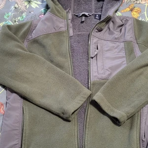 Grön fleecejacka från Lands' End - Säljer en grön fleecejacka/ tjock tröja från Lands' End med dragkedja och huva. Jackan har en praktisk bröstficka och är perfekt för kyliga dagar. Den är mjuk och bekväm med långärmade ärmar. (Det finns en hund hemma.)