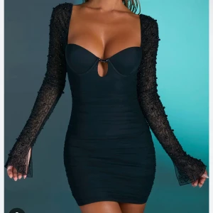 Svart klänning med pärldetaljer - Snygg svart kortklänning med långa ärmar och pärldetaljer. Klänningen har en djup v-ringning och en liten öppning framtill. Den är tight och har en knytning i ryggen för extra stilpoäng.