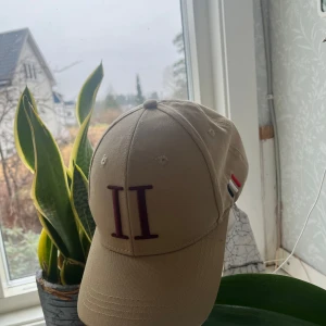 Beige keps från Les Deux - Snygg beige keps från Les Deux med broderad logga i vinrött framtill. Kepsen har en justerbar rem baktill för perfekt passform. Tillverkad i bomull och designad i Köpenhamn. Perfekt för en stilren look.