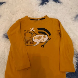 Gul långärmad tröja med tryck - Säljer en gul långärmad tröja från Basic Needs by Name It. Tröjan har ett coolt tryck med texten 'Hello I'm Cool' och motiv av en telefon och verktyg. Perfekt för barn som gillar att sticka ut med sin stil!