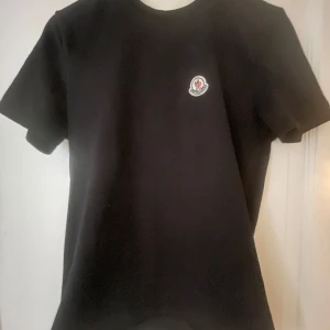 Svart t-shirt från Moncler - Säljer en stilren svart t-shirt från Moncler med en liten logotyp på bröstet. Perfekt för en avslappnad och trendig look. T-shirten har korta ärmar och är tillverkad i ett mjukt material. Oäkta kan diskutera pris självklart 😁