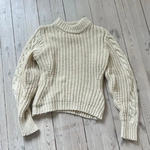 Beige stickad tröja - Mysig beige stickad tröja med kabelstickade detaljer på ärmarna. Tröjan har en hög krage och långa ärmar, perfekt för kyligare dagar. Den ribbade strukturen ger en klassisk look.