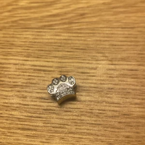 Silverfärgad berlock i form av tass - Charmig berlock i form av en tass, dekorerad med glittrande stenar. Perfekt för att fästa på ett armband eller halsband för en personlig touch. Den silverfärgade ytan ger en elegant och stilren look.