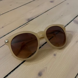 Beige solglasögon med bruna linser - Snygga solglasögon i beige med bruna linser. Ramen har en klassisk rundad form och små dekorativa detaljer vid sidorna. Perfekta för en solig dag och ger en stilren look. Använda fåtal gånger men fortfarande i mycket bra skick ocj utan defekter. Pris kan diskuteras!❤️