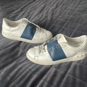 Valentino sneakers i vitt och blått - Snygga Valentino sneakers i vitt med en bred blå rem över ovansidan. Skorna har snörning och en klassisk design med en bekväm sula. Perfekta för en stilren look.