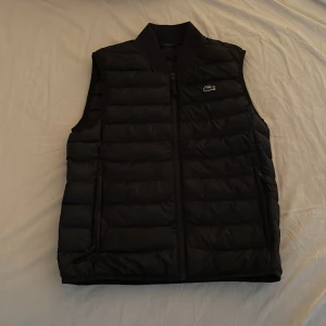 Svart dunväst från Lacoste - Säljer en stilren svart dunväst från Lacoste med dragkedja framtill. Västen har en quiltad design och en liten logotyp på bröstet. Perfekt för lager-på-lager under kyligare dagar.