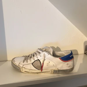 Vita sneakers från Philippe Model - Snygga vita sneakers från Philippe Model med grå detaljer och blå häl. Skorna har snörning och en röd detalj på sidan. Perfekta för en avslappnad stil. Nypris: 3500kr                                Slitna men fortfarande stiliga 