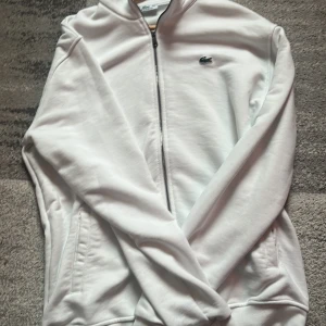 Vit zip-tröja från Lacoste - Säljer en stilren vit zip-tröja från Lacoste med det klassiska krokodilmärket på bröstet. Tröjan har långa ärmar och dragkedja framtill. Perfekt för en avslappnad look.Storleken är liten på grund av tvätt och sitter väldigt bra.Pris kan diskuteras