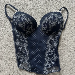 Mörkblå korsettopp med spetsdetaljer - Elegant mörkblå korsettopp med vita prickar och vackra spetsdetaljer. Den har justerbara axelband och en hakknäppning bak. Perfekt för en stilfull look. Står ej storlek men skulle gissa på M-L. Inga skavanker!