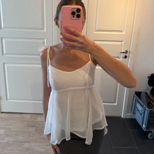 Vit ärmlös blus - Säljer en söt vit blus med tunna axelband och knytning på axlarna. Blusen har en luftig passform och är perfekt för varma dagar. Den har en lätt och fladdrig design som ger en romantisk känsla.