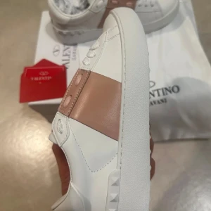 Valentino sneakers med rosa detalj - Snygga vita Valentino sneakers med en bred rosa rem över snörningen. Skorna har en stilren design med diskreta nitar på sidorna och en mönstrad sula för bra grepp. Perfekta för en trendig look.
