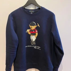 Mörkblå sweatshirt från Ralph Lauren - Säljer en mörkblå sweatshirt från Ralph Lauren med en ikonisk Polo Bear-design på framsidan. Tröjan har långa ärmar och rund halsringning. 🐻