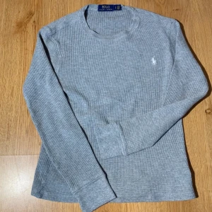 Grå långärmad tröja från Ralph Lauren - Säljer en stilren grå långärmad tröja från Polo Ralph Lauren. Tröjan har ett diskret rutmönster och det klassiska broderade logotypen på bröstet. Perfekt för en avslappnad och snygg look.