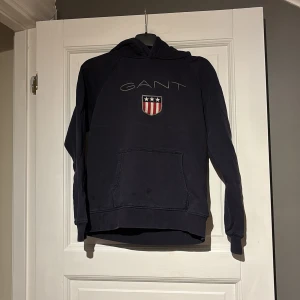 Svart hoodie från GANT - Snygg svart hoodie från GANT med ett stort tryck av märkesnamnet och en sköld med stjärnor och ränder på framsidan. Tröjan har en klassisk känguruficka och långa ärmar. Perfekt för en avslappnad stil.