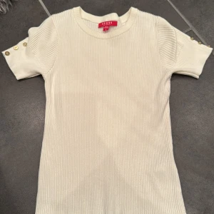 Vit ribbad topp från Guess - Säljer en stilren vit ribbad topp från Guess med korta ärmar och guld knappar på axlarna. aldrig blivit använd.