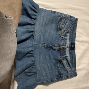 Blå jeanskjol med volang - Snygg blå jeanskjol med volangdetaljer. En bekväm passform. Perfekt för en avslappnad stil med en twist. Perfekt till sommaren. Har även en slits på sidan