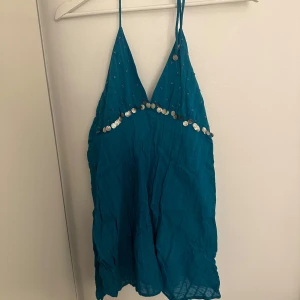Turkos klänning med paljetter - Säljer en turkos klänning med smala axelband och dekorativa paljetter längs bysten. Klänningen har en v-ringad design och snörning i ryggen för en justerbar passform. Perfekt för sommarens festligheter! 