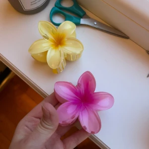 Hårklämmor i form av blommor - Två hårklämmor i form av blommor, en i rosa och en i gul. De har en glansig yta och är formade som plumeriablommor, vilket ger en söt och somrig känsla. Perfekta för att sätta upp håret med stil. Oanvända 
