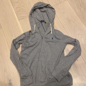 Grå hoodie från Polo Ralph Lauren - Säljer en stilren grå hoodie från Polo Ralph Lauren med dragkedja och en liten blå logga på bröstet. Tröjan har långa ärmar och en praktisk huva med snören. Perfekt för en avslappnad look. Nypris 1800, slutsåld överallt.