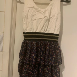 Blommig ärmlös klänning med volang - Klänning med vit ärmlös överdel och blommig kjol i mörkblått. Den har en randig midja i svart och guld. Kjolen har volanger. Finns ingen tagga med storlek men jag antar att det är en S. Hör av dig vid frågor!😊