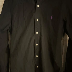 Svart skjorta från Polo Ralph Lauren - Säljer en stilren svart skjorta från Polo Ralph Lauren i strlk:S. Skjortan har långa ärmar och knappar fram. Den är i bomull och har en liten lila logga broderad på bröstet. Perfekt för både vardag och fest! Skicket är mycket bra använd ett fåtal gånger. Pris kan diskuteras vid snabb affär!🔥🔥🔥