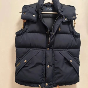 Mörkblå dunväst från Polo Ralph Lauren - Säljer en snygg mörkblå dunväst från Polo Ralph Lauren. Västen har en dragkedja och knappar framtill, samt praktiska fickor. Perfekt för höst och vinter, håller dig varm och stilren. Passar både till vardags och lite finare tillfällen.