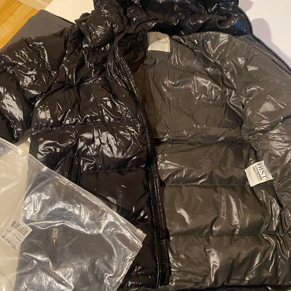 Säljer en snygg svart dunjacka från Moncler i nytt skick. Jackan har en glansig finish och är perfekt för vintern. Den har en dragkedja framtill och en praktisk huva. Moncler-loggan syns på ärmen och den kommer med originalpåse. Strl 4/M. Jackor.