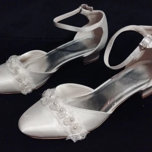 Vita satinklackskor med pärldetaljer - Säljer ett par eleganta vita skor i satin med en liten klack. Skorna är dekorerade med pärlor och spetsdetaljer framtill. De har en justerbar rem runt ankeln för bättre passform. Normal i storleken. Perfekta för bröllop eller bal!