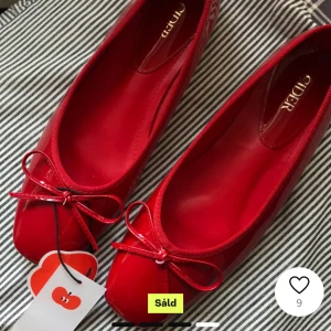 Röda ballerinaskor med rosett - Säljer ett par snygga röda ballerinaskor i syntetmaterial. De har en söt rosett på ovansidan och är perfekta för både vardag och fest. Skorna är i nytt skick och har en glansig finish som verkligen sticker ut. Perfekta för att lägga till en färgklick i din outfit!