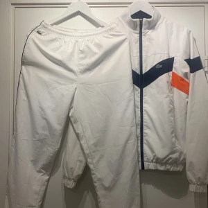 Vit vindjacka och byxor från Lacoste - Säljer en snygg vit vindjacka och vindbyxor från Lacoste med coola marinblå och orange detaljer. Jackan har en dragkedja framtill och elastiska muddar vid ärmslut och nederkant. Perfekt för träning 🧡💙