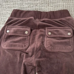 Bruna mjukisbyxor från Juicy Couture - Säljer ett par bruna mjukisbyxor från Juicy Couture i storlek XXS. Byxorna har en loose passform med breda ben och är tillverkade i mjuk sammet. De har praktiska fickor med knappar bak och en bekväm resårmidja. Perfekta för en avslappnad stil!