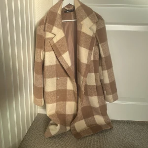 Rutig kappa från Gina Tricot - Säljer en superfin rutig kappa från Gina Tricot i beige och brun. Den har en klassisk design med stora rutor och knappar framtill. Perfekt för höst och vår, och ger en stilren look. Kappan är i mycket bra skick och passar perfekt för att hålla sig varm och snygg.