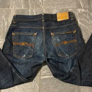 Säljer ett par mörkblå jeans från Nudie Jeans i bra skick. De har en klassisk femficksdesign med orange sömmar och en liten reva vid grenen. Perfekta för en avslappnad stil. Passar både till vardags och fest! Pris ej hugget i sten 