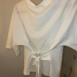 Vit topp med en rosett från Nelly🎀 - Säljer en stilren vit topp från Nelly i storlek L. Den har korta ärmar och en snygg knytning bak på ryggen som ger en fin siluett. Perfekt för både vardag och festliga tillfällen. Toppen är i bra skick och redo för en ny garderob!