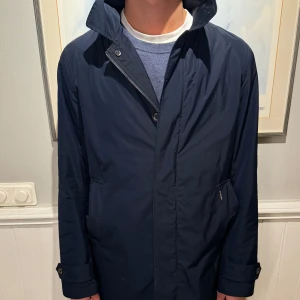 Mörkblå Ben Sherman rock - Säljer en stilren mörkblå rock i mycket bra skick. Den har en klassisk design med dragkedja och knappar framtill. Perfekt för höst och vår och vinter, med långa ärmar och en snygg krage. Rocken är både praktisk och elegant, idealisk för både vardag och fest.