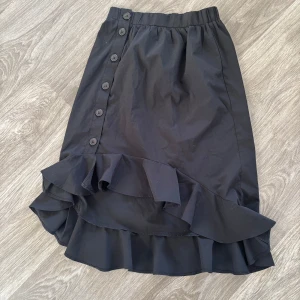 Svart kjol med volang och knappar - Snygg svart kjol med volangdetalj längst ner och knappar längs sidan. Perfekt för både vardag och fest! Den har en elastisk midja för bekväm passform. Kjolen är i ett lätt och luftigt material, vilket gör den idealisk för vår och sommar.