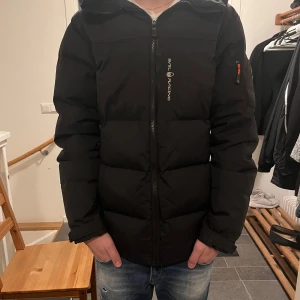 Svart pufferjacka från Sail Racing - Säljer en svart pufferjacka från Sail Racing i mycket bra skick. Jackan är använd en vinter men har inga defekter. Jackan har en dragkedja framtill och en cool patch på ärmen. Jackan är lite längre där bak då de ska vara Rump värme, men man kan dra upp den som ni kan se att jag gjorde på 3de bilden. Modellen är 180cm lång. Köpt för 3500. 