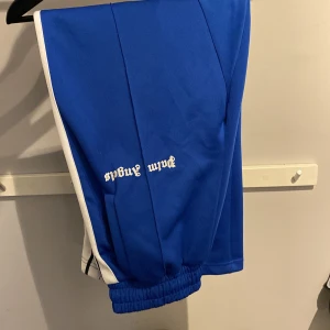 Blå byxor från Palm Angels - Säljer ett par snygga blå byxor från Palm Angels. De har en vit rand längs sidorna och en dragkedja vid bensluten. Byxorna är i mycket bra skick och perfekta för både träning och vardag. Elastisk midja för extra komfort. Passar perfekt för dig som vill ha en sportig och trendig stil! Skick 9,5/10, andvända fåtal gånger. Pris kan diskuteras 