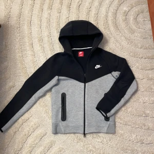 Nike tech fleece - Säljer en snygg och bekväm huvtröja från Nike tech  i grått och svart. Den har en dragkedja framtill och en praktisk huva. Perfekt för både träning och vardag. Tröjan är i mycket bra skick och gjord av mjukt material för extra komfort. Väldigt bra skick. (Ny version)