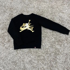 Svart Nike Air Jordan tröja - Säljer en snygg svart Nike Air Jordan tröja. Den har ett coolt guldtryck på framsidan och texten 'Air Jordan' på sidan. Perfekt för både vardag och träning. Tröjan är i mycket bra skick och redo för nya äventyr!