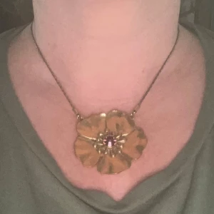 Guldigt halsband med blomma från Pilgrim - Säljer ett halsband från Pilgrim med en stor blomma i guldton. Blomman har en vacker lila sten i mitten som ger en fin kontrast. Perfekt för att piffa upp en outfit till fest eller speciella tillfällen. Kedjan är tunn och elegant, vilket gör att blomman verkligen står ut. Ett måste för smyckesälskare! 🌸✨