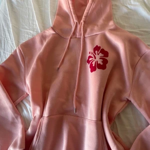 Rosa hoodie med hibiskusmotiv - Säljer en superfin rosa hoodie med en stor röd hibiskusblomma på bröstet och texten 'Ocean Beach, California' på ryggen. Perfekt för chill dagar eller när du vill ha en touch av sommar året runt. Den har en skön passform och en praktisk ficka framtill. Huvan har dragsko för justering. 🏄‍♀️🌺