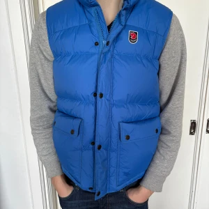 Blå dunväst från Fjällräven - Säljer en blå dunväst från Fjällräven, Expedition Series 1974. Västen har en quiltad design med knappar och två stora fickor framtill. Perfekt för kyliga dagar och håller dig varm med stil. Passar bra för både höst och vinter.