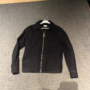 Marinblå overshirt från Weekday - Säljer en stilren marinblå overshirt från Weekday i storlek S. Jackan har en praktisk dragkedja framtill och en enkel bröstficka. Perfekt för höst och vår, med en klassisk krage och långa ärmar. Passar både till vardags och lite mer uppklädda tillfällen.