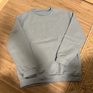 Ljusblå sweatshirt med 'Malibu' tryck - Säljer en supermjuk ljusblå sweatshirt med 'Malibu' tryck på framsidan. Tröjan har långa ärmar och är perfekt för en avslappnad stil. Den är i bra skick och gjord av ett bekvämt material som känns skönt mot huden.